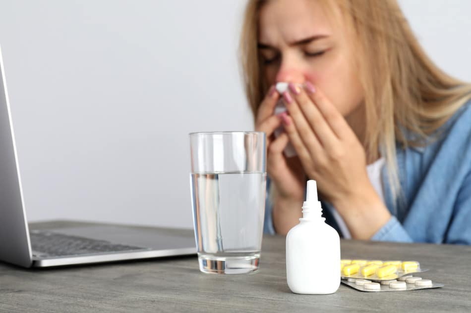fotografía de un medicamento para el flujo nasal en primer plano junto a un vaso, en segundo plano una mujer sonándose la nariz.
